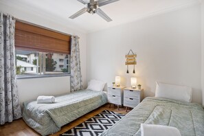 Ein Schlafzimmer mit zwei Betten, einem Deckenventilator, einem Fenster mit Vorhängen, Nachttischen mit Lampen und einer Wanddekoration.