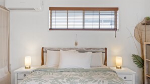 A bedroom with a bed, two bedside tables with lamps, a window with blinds, and an air conditioning unit.