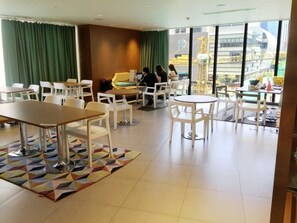 Un hall d’hôtel moderne doté de plusieurs espaces de détente, comprenant des tables et des chaises, et de grandes fenêtres offrant une vue sur le paysage urbain.