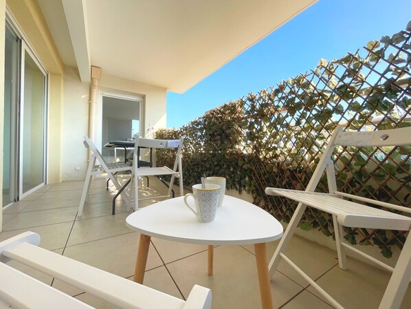 A balcony with a white table, two white chairs, a white bench, and a white fence with greenery.