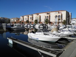 A marina with several boats docked, a wooden dock, and a building with multiple balconies in the background.