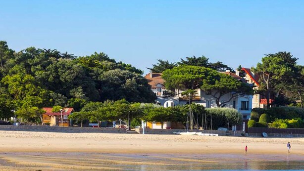 Ein Strand mit sandigem Ufer, wenigen Personen und Gebäuden mit roten Dächern im Hintergrund.