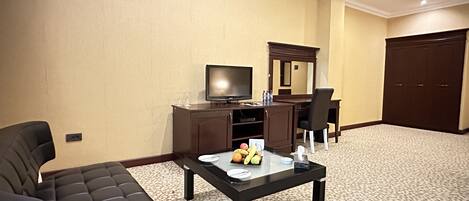 Habitación de hotel con sofá de cuero negro, una mesa de centro de vidrio con frutas, un televisor, un escritorio y un armario.