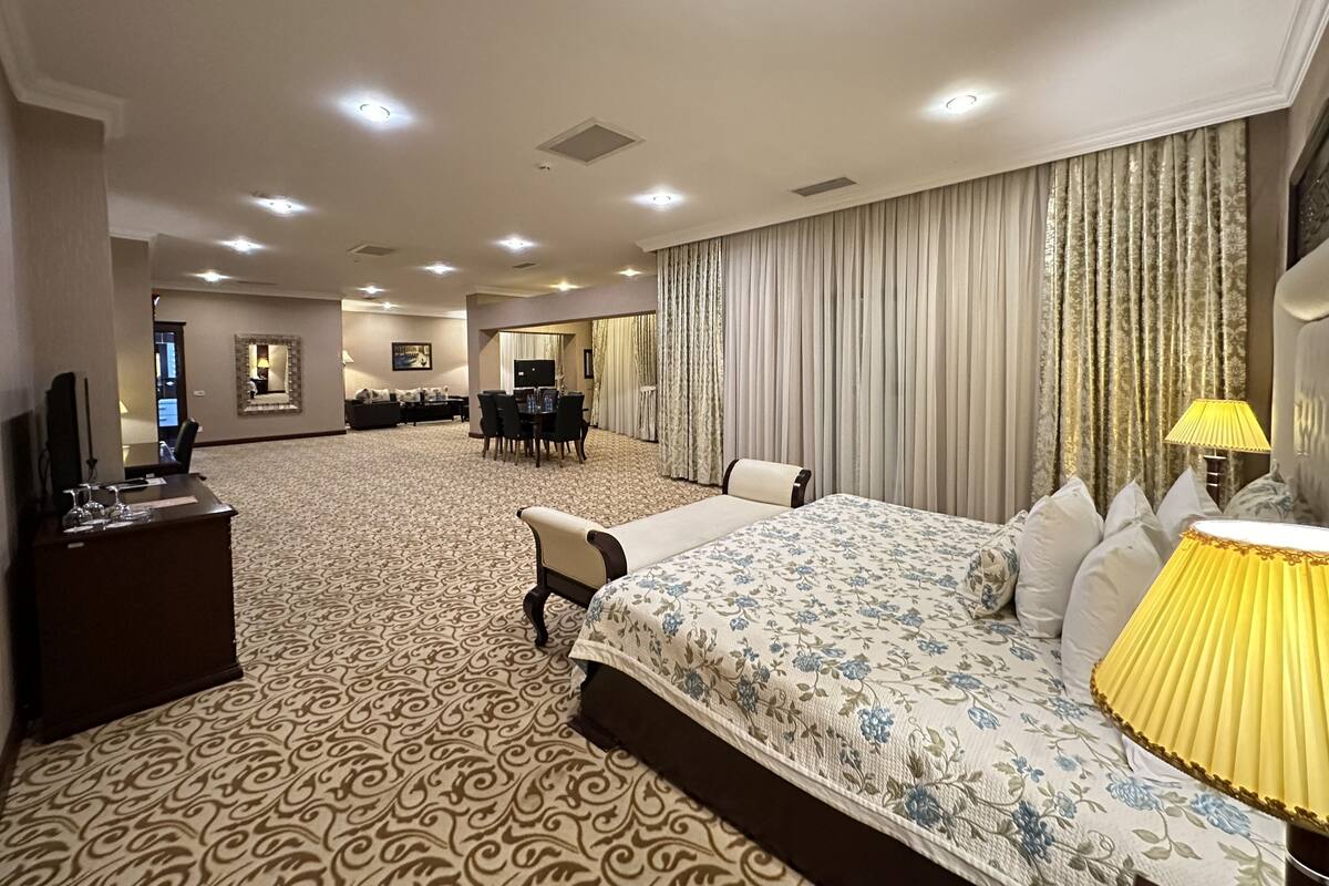 A hotel room with a large bed, a desk, a TV, and a dining area in the background.
