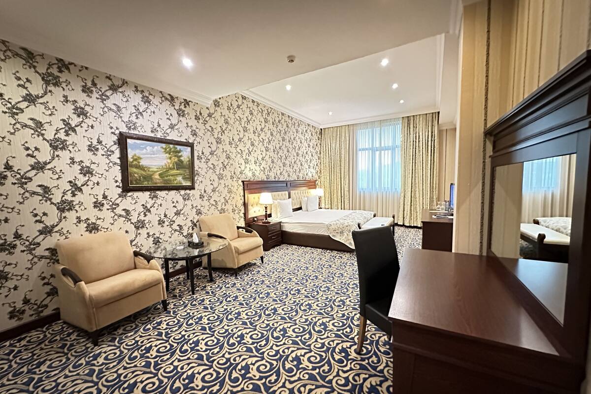 A hotel room with a bed, a desk, two armchairs, a small table, and a large mirror.