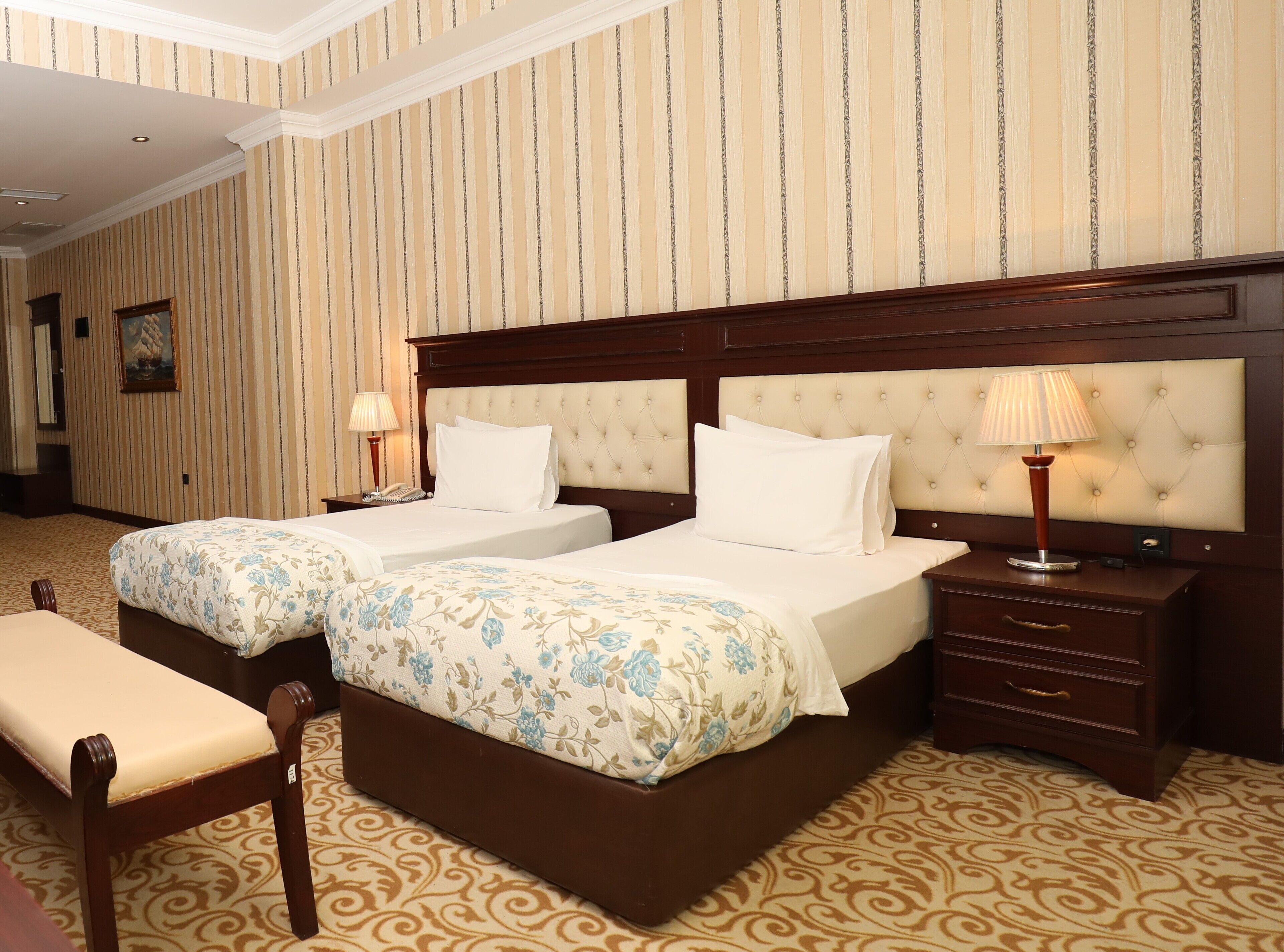 A hotel room with two beds, a wooden headboard, a bedside table with a lamp, and a patterned carpet.