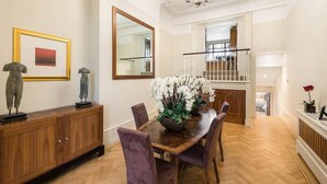 Un coin repas avec une table en bois, des chaises violettes et un vase rempli de fleurs blanches. Un grand miroir et une œuvre d’art encadrée ornent le mur.