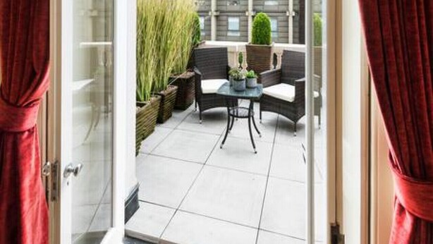 Une porte ouverte donnant sur un balcon avec vue sur le clocher d’une église, un espace extérieur aménagé avec des sièges et des plantes en pot.