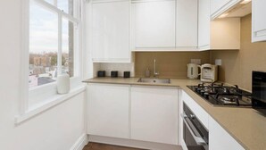A modern kitchen with white cabinets, a gas stove, a sink, and a window overlooking a cityscape. - Chelsea Apartment | King`s Road, River Thames & Thames Clipper (London)