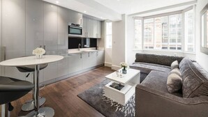 A modern kitchen with a built-in oven and microwave, a dining area with a round table and bar stools, a gray sofa, and a large window offering a city view. - Chelsea Apartment | King`s Road, Sloane Sq, Lift & Underground Parking (London)