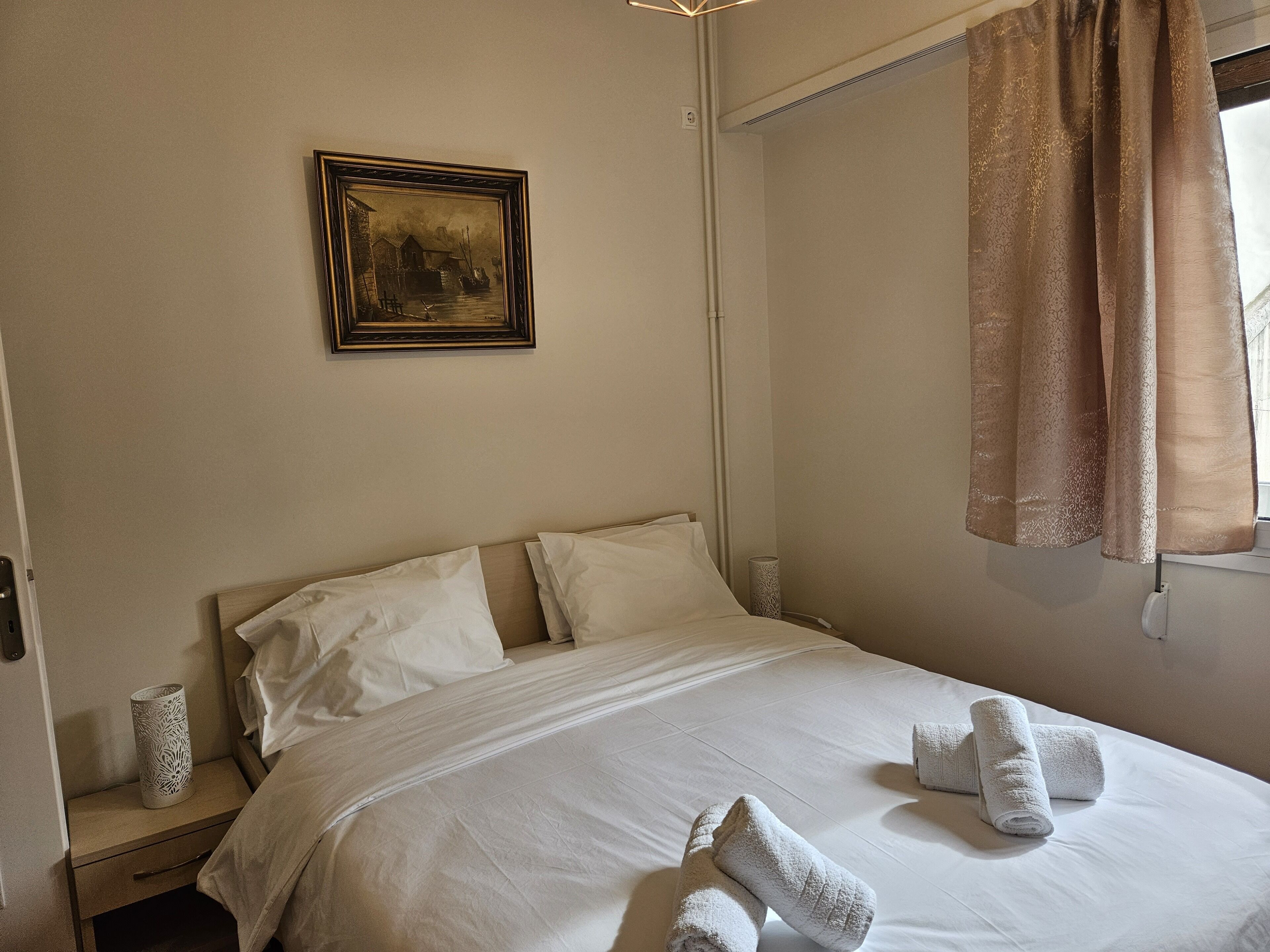 A neatly made bed with white linens and two rolled towels, a framed picture on the wall, and a window with curtains.