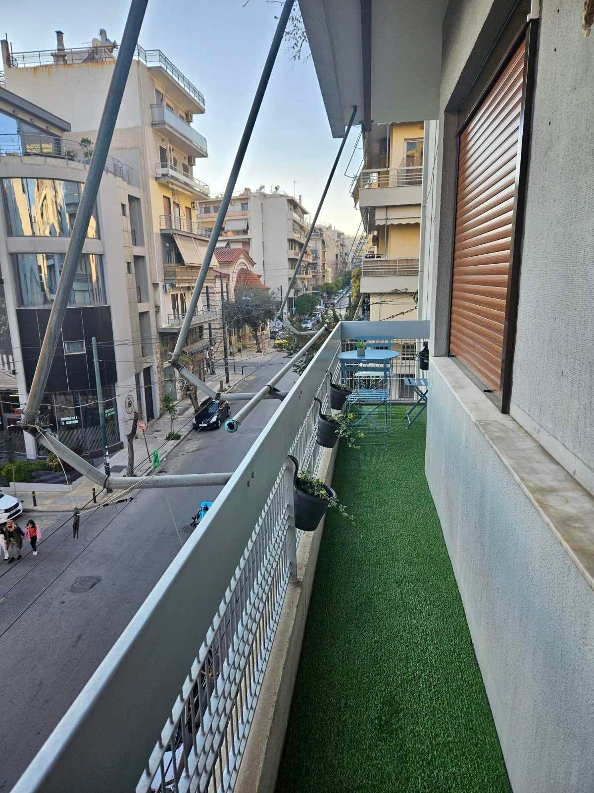 A balcony with artificial grass, a metal railing, and a view of a street with buildings and parked cars.