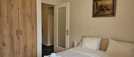 A bedroom with a large bed, a wooden wardrobe, a painting on the wall, and a door leading to another room.