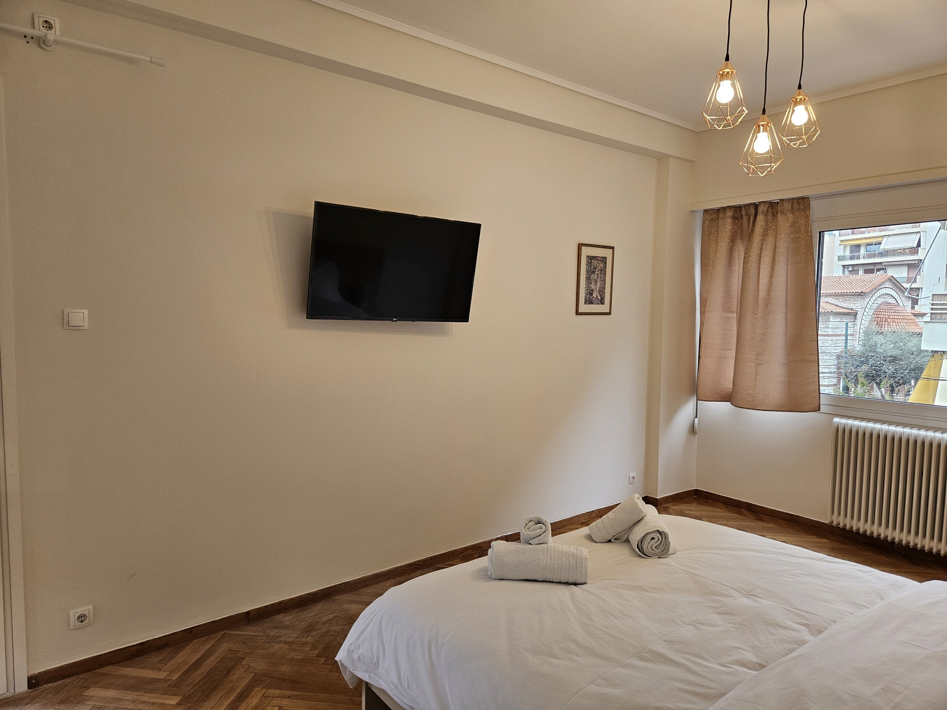 A bedroom with a bed, a flat-screen TV mounted on the wall, a framed picture, and a window with curtains.