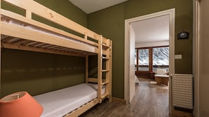 Ein Schlafraum mit Etagenbetten, einer Holzleiter, einem kleinen Tisch mit Lampe und Blick auf eine verschneite Landschaft durch ein Fenster.