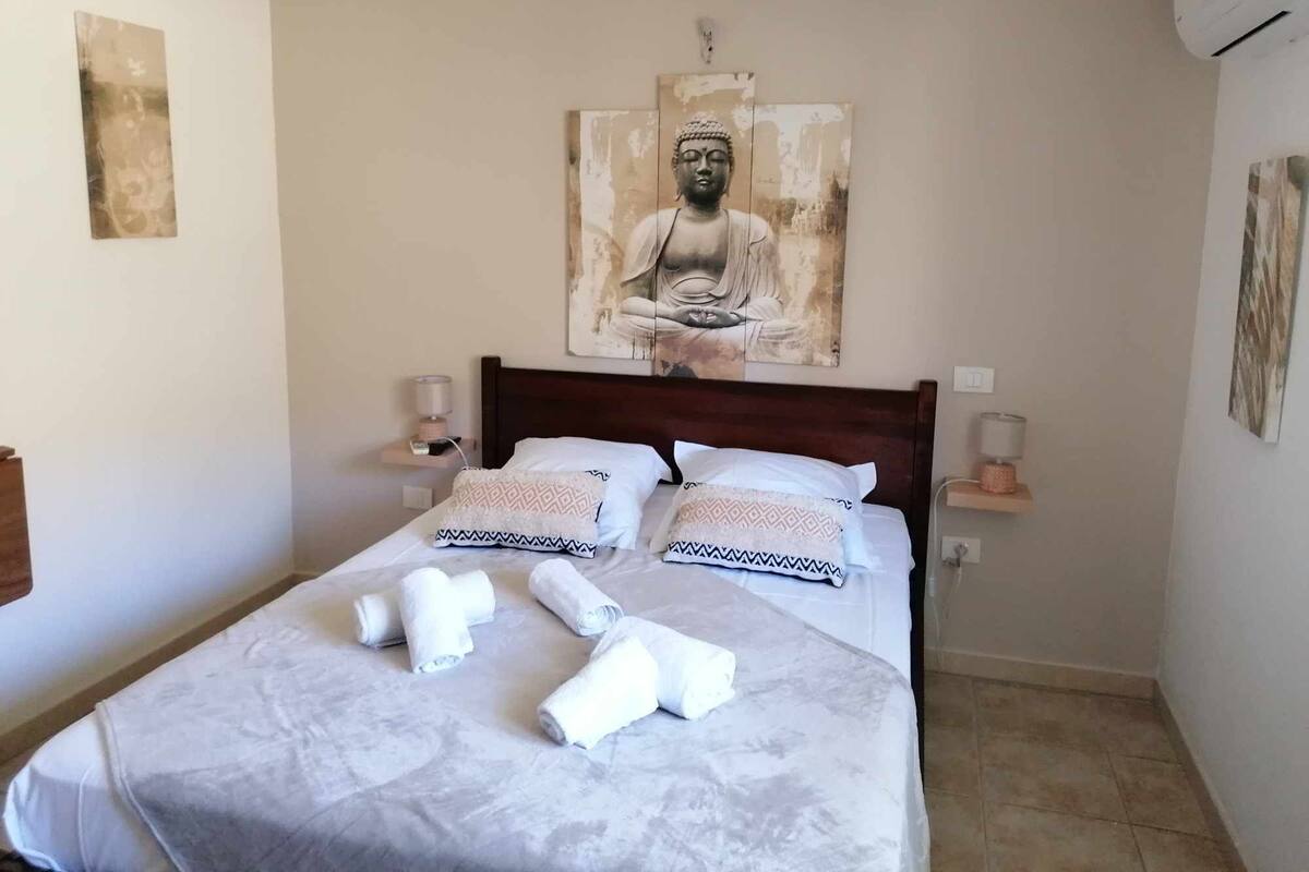 Un lit bien fait, avec des serviettes, une tête de lit en bois et un grand tableau représentant Bouddha au-dessus.