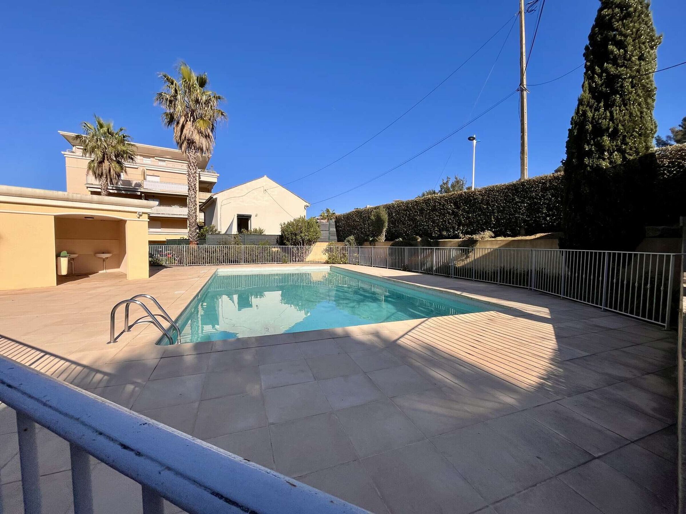 Piscina com deck revestido, cercada por uma grade, adjacente a um edifício com varanda.
