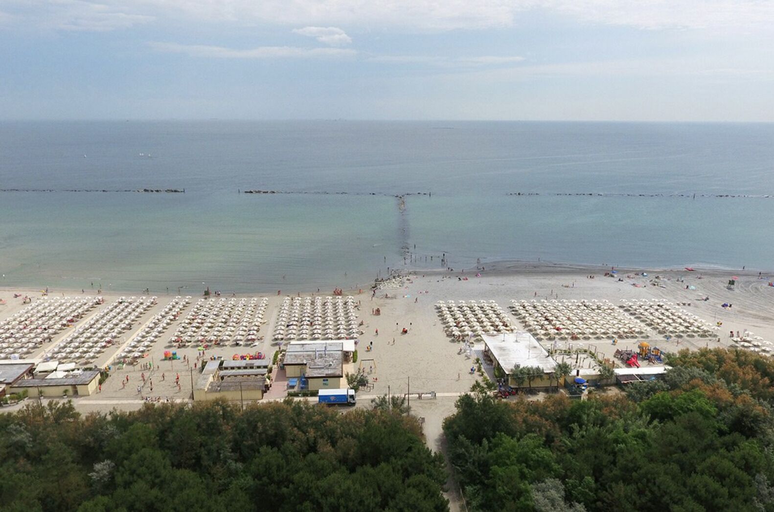 A beach with rows of umbrellas and beach chairs, a pier extending into the sea, and a parking area with a few vehicles.