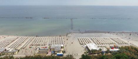 A beach with rows of umbrellas and beach chairs, a pier extending into the sea, and a parking area with a few vehicles.
