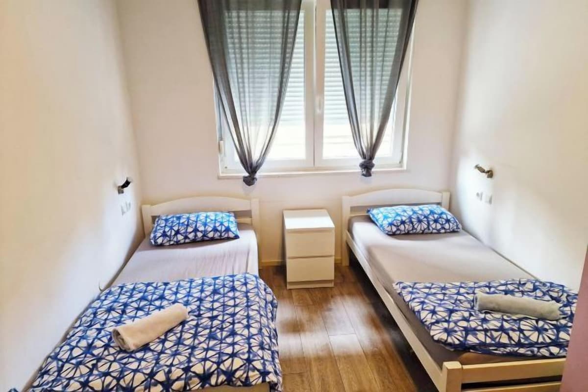 Una habitación pequeña y limpia con dos camas individuales, una mesita de noche y un ventanal con cortinas.