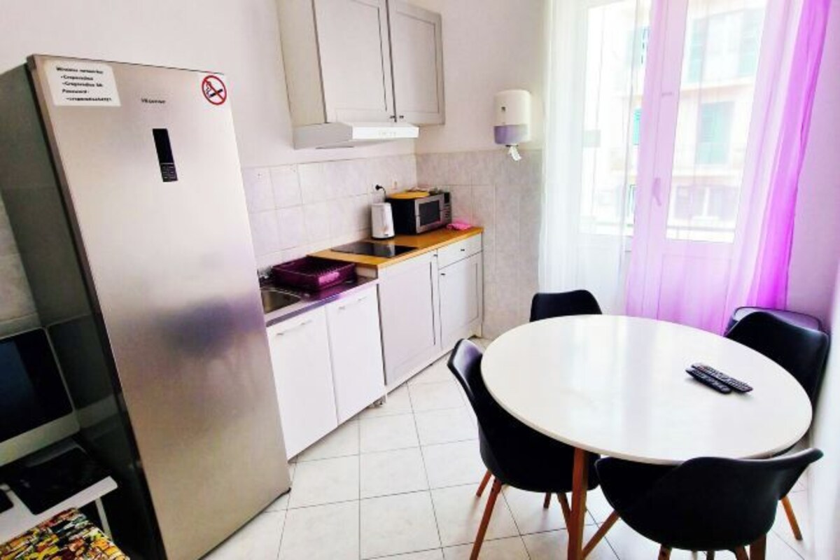 Een praktische keuken met een koelkast, magnetron en een klein aanrecht. Er is een eettafel met stoelen en een raam met gordijnen.