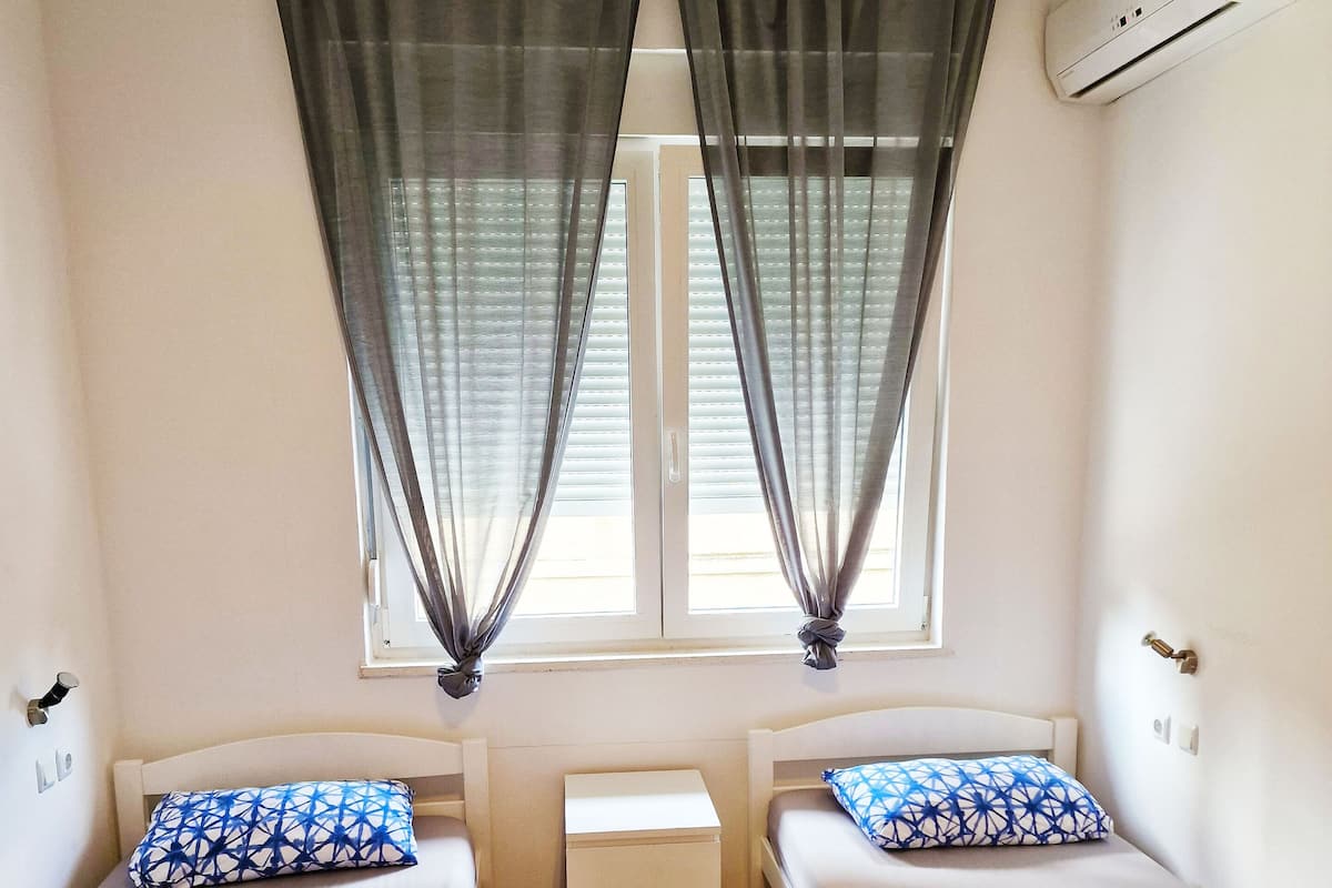 A room with two beds, a small bedside table, a window with curtains, and an air conditioning unit.
