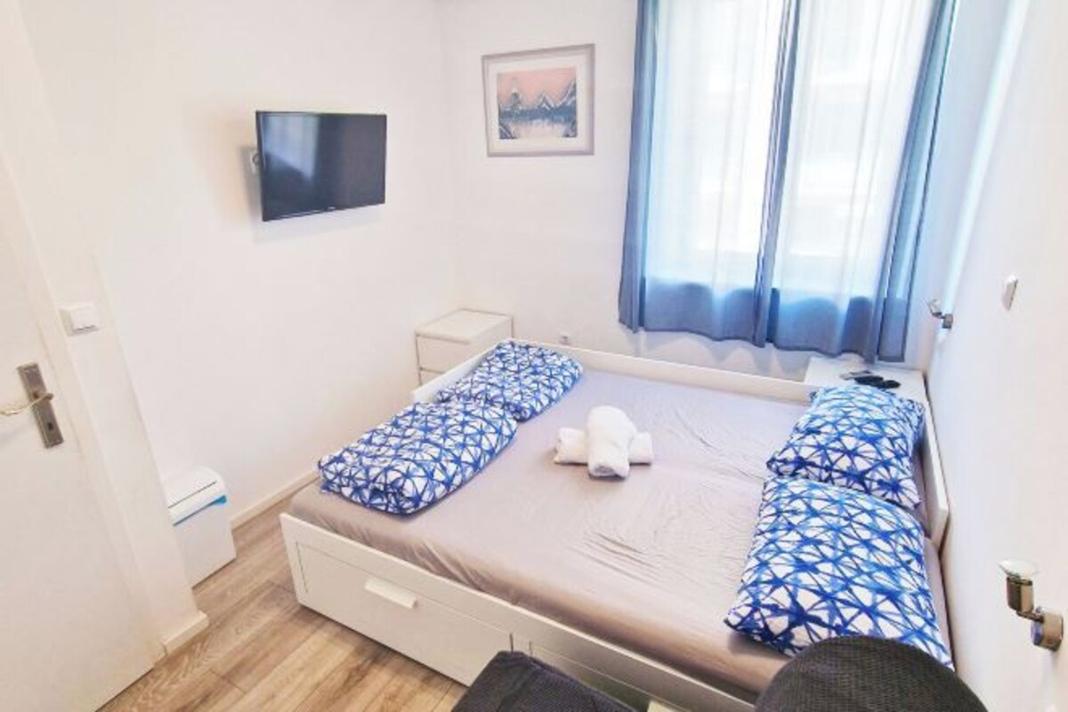 A bedroom with a bed, a TV mounted on the wall, a small bedside table, a window with curtains, and a picture on the wall.