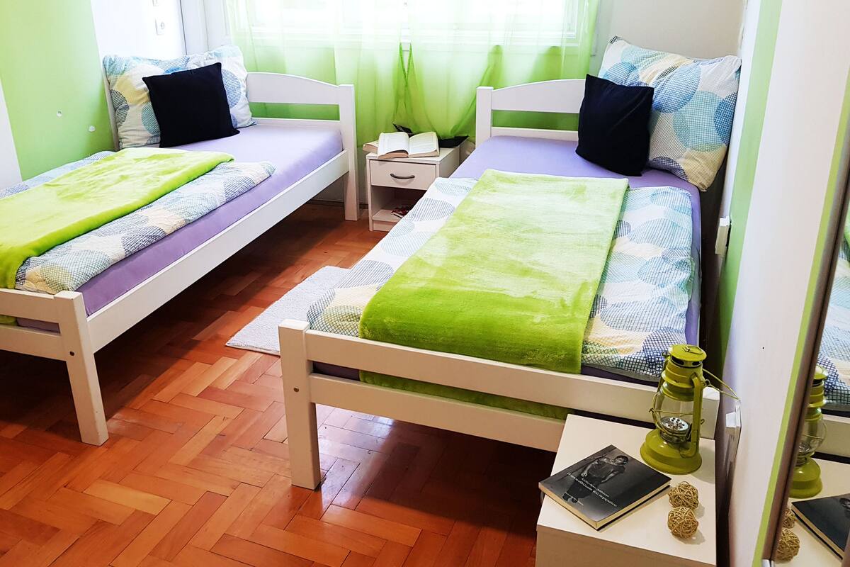 A room with two single beds, each with a green blanket and a pillow, a bedside table with a book and a bottle, and a small table with a book and decorative items.
