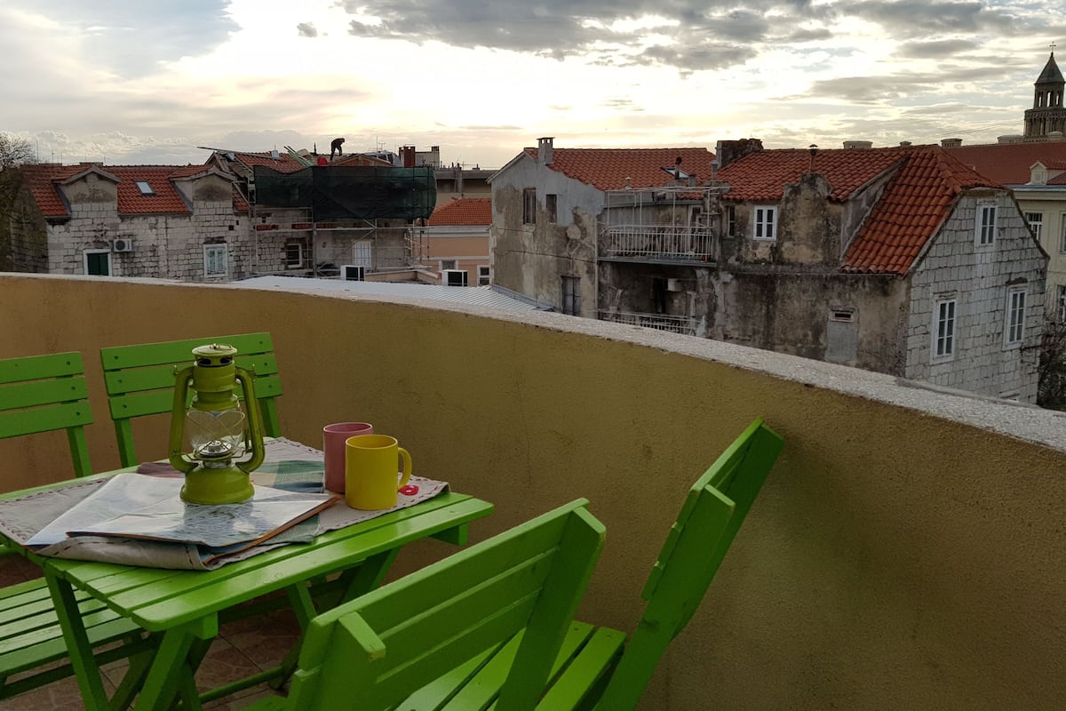 A balcony with green chairs and a table, overlooking a town with red-roofed buildings and a church tower.