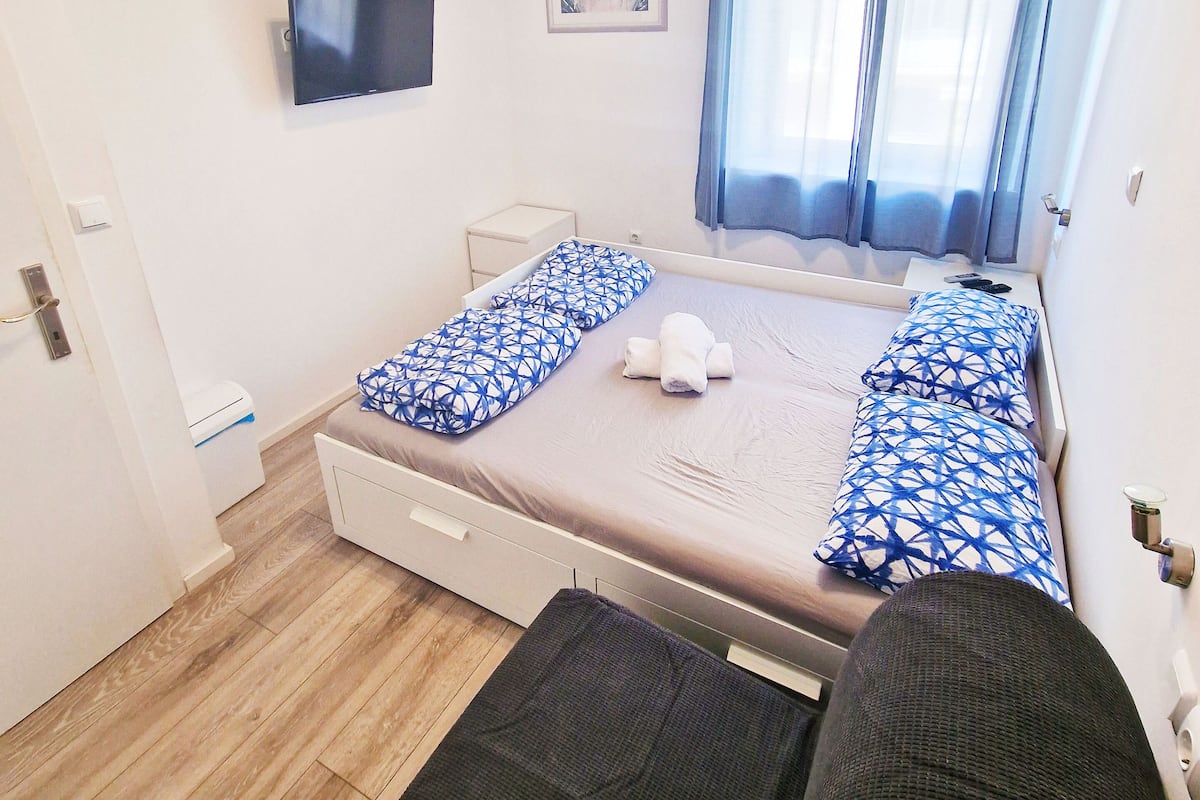 A bedroom with a bed, a TV, a window with curtains, a small bedside table, and a wall-mounted air conditioner.