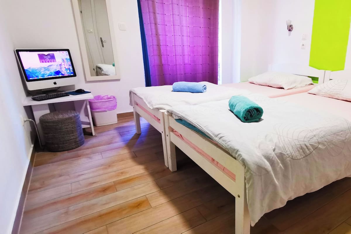 A bedroom with two beds, a desk with a computer, a trash bin, and a wall-mounted light fixture.