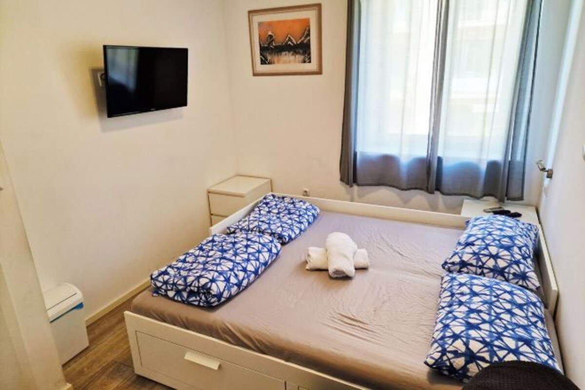 Een eenpersoonskamer met een flatscreen televisie aan de muur, een klein nachtkastje, een raam met gordijnen en een schilderij boven het bed.