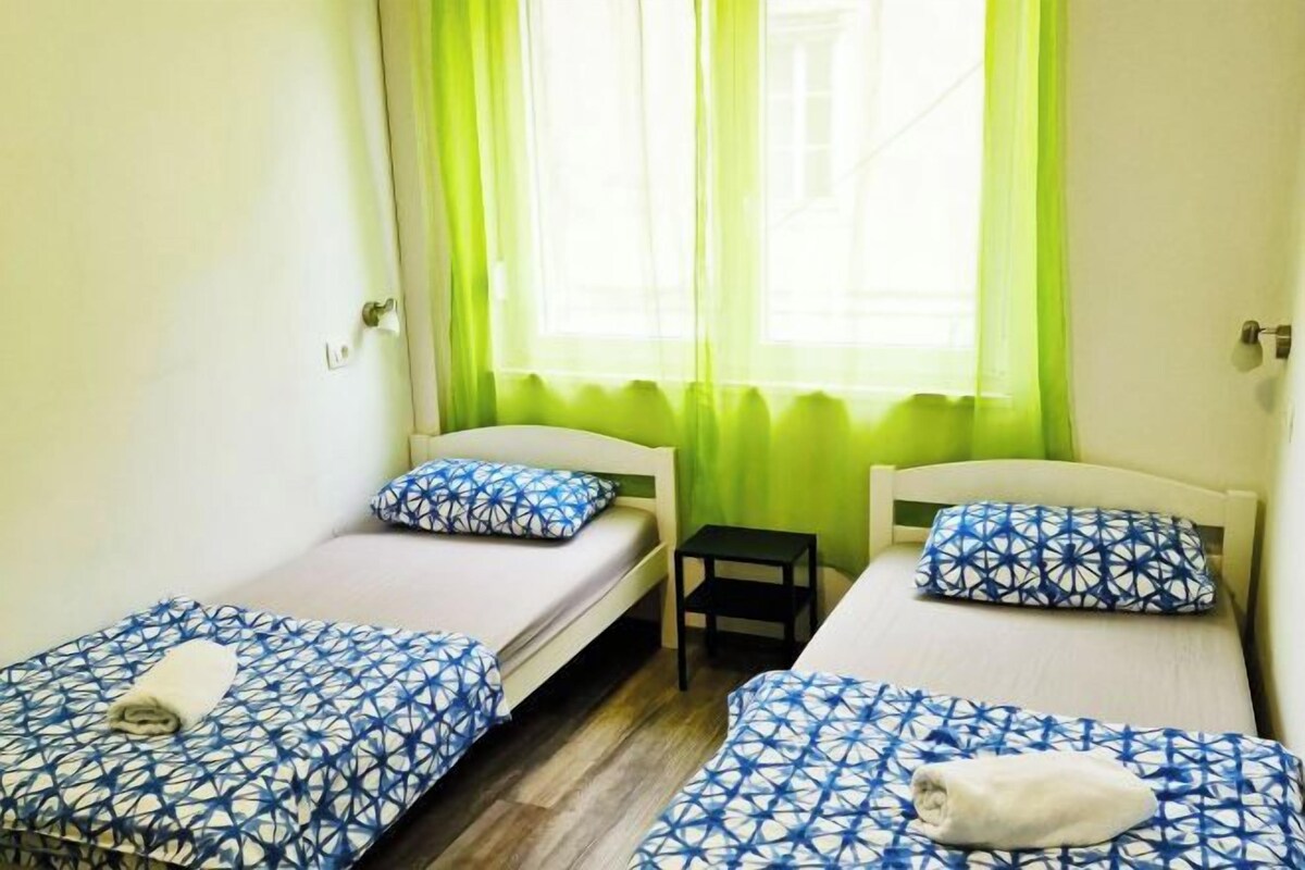 Quarto com duas camas de solteiro, cada uma com colcha estampada e almofadas combinando, uma pequena mesa de cabeceira e uma janela com cortinas verdes.
