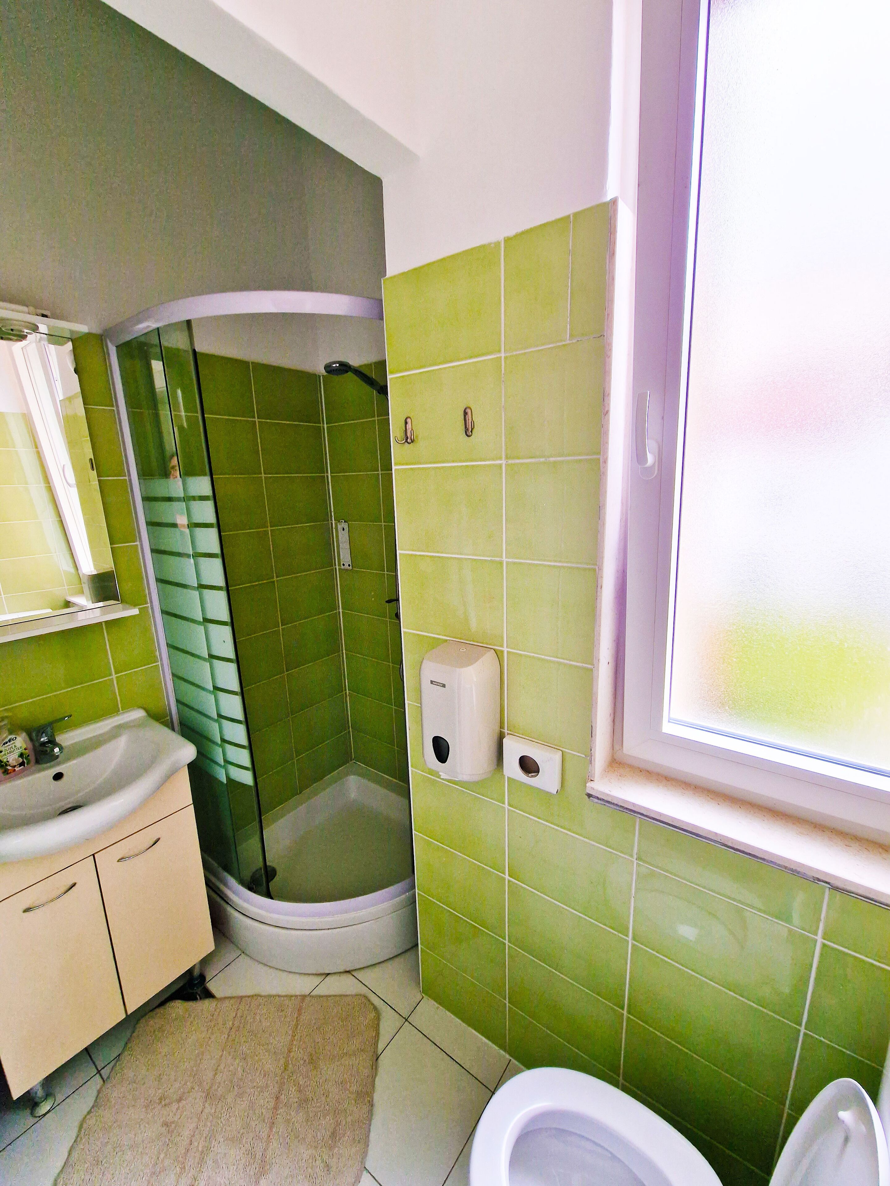 Casa de banho com azulejos verdes, uma pia, uma sanita e uma janela.