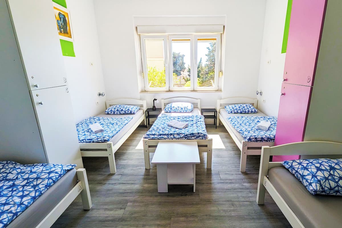 A dormitory room with four single beds, a central bench, and a window with a view of trees.