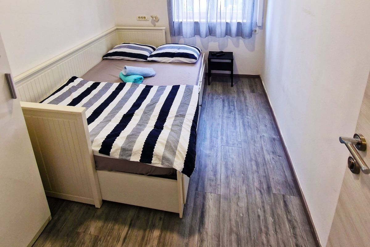 A small, narrow hotel room with a single bed, a bedside table, a TV mounted on the wall, and a window with sheer curtains.