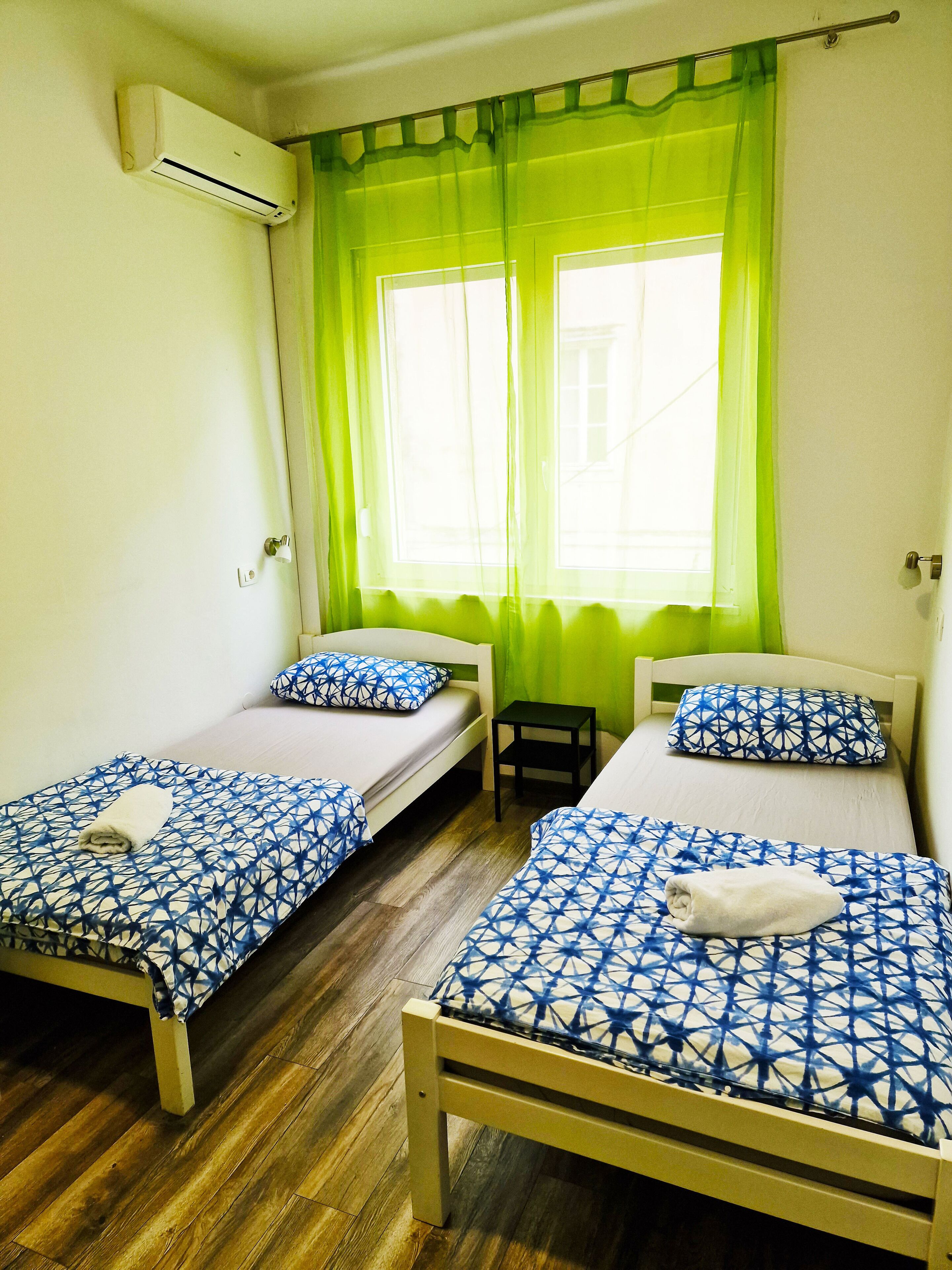 A room with two single beds, a small table, a window with green curtains, and an air conditioning unit on the wall.