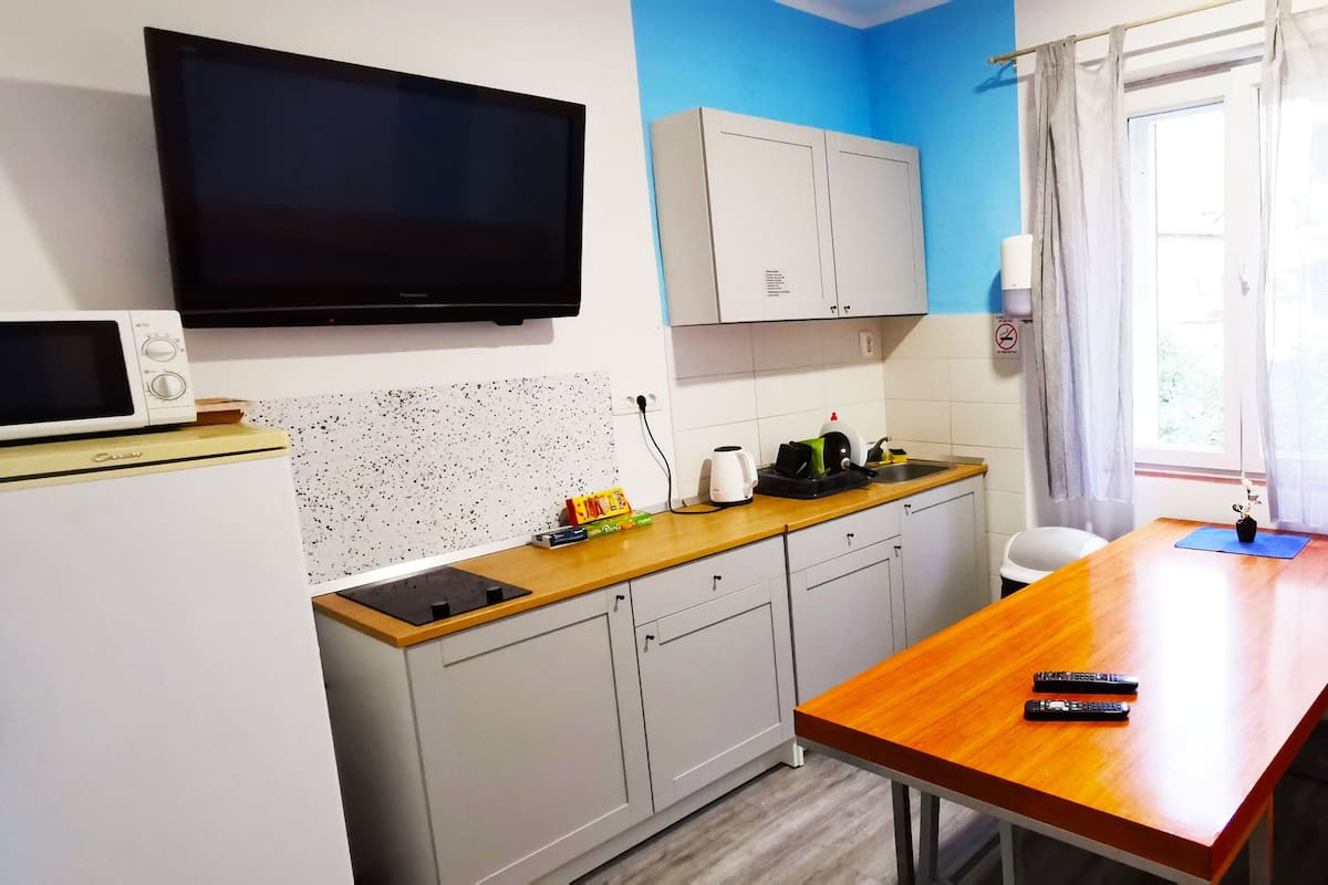 A compact kitchen with a microwave, kettle, and toaster on the counter, a wooden table, and a TV mounted on the wall.