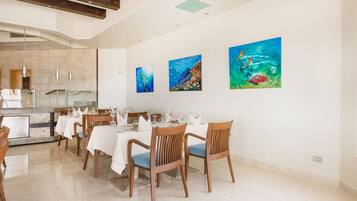 A dining area with wooden chairs and tables, a kitchen in the background, and three paintings on the wall.