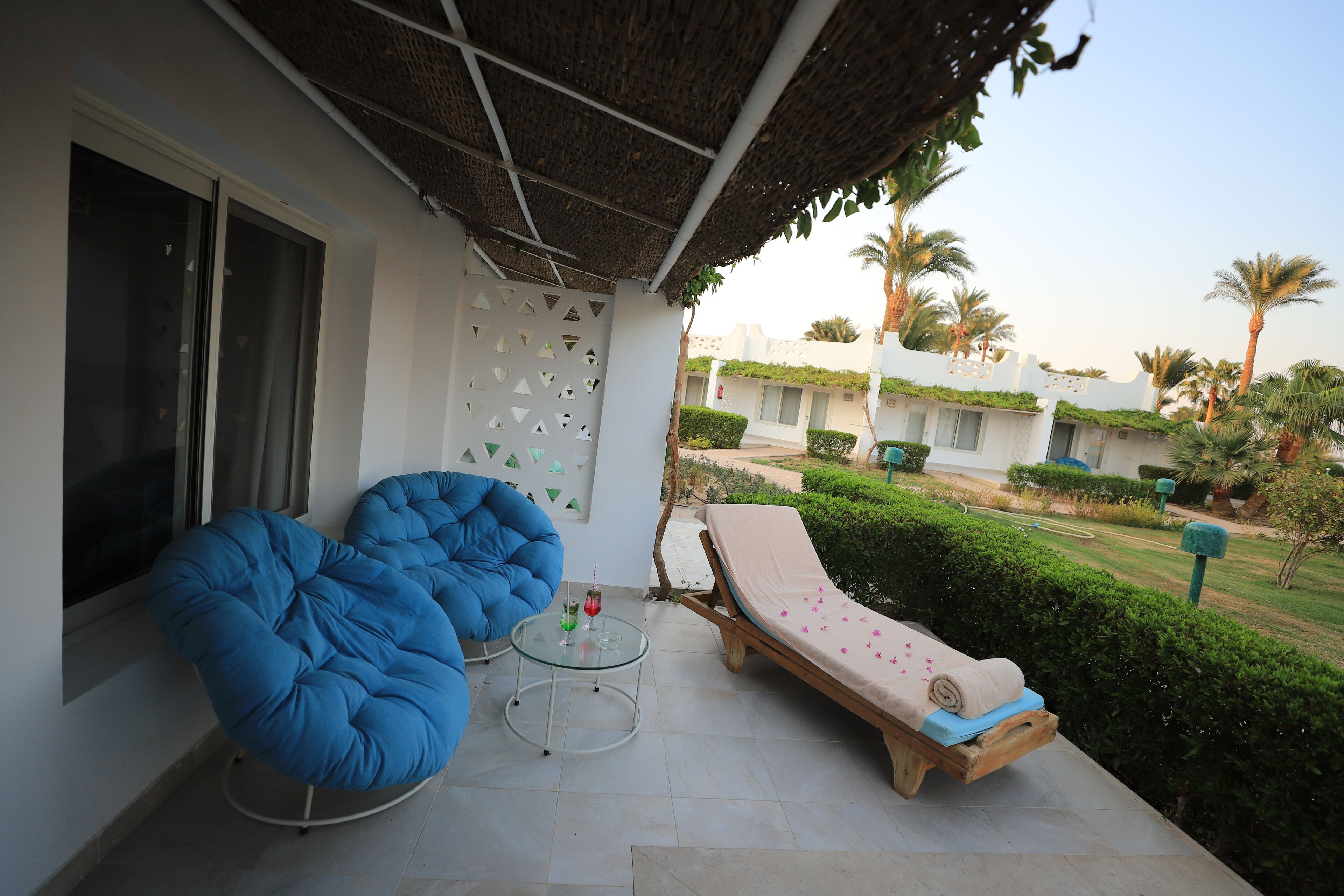 Un área de estar al aire libre techada con una silla de lounge, un sillón informal tipo bean bag azul y una pequeña mesa con una bebida.