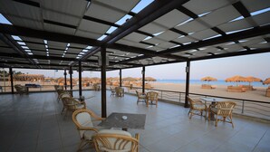Un espacio de estar al aire libre cubierto, con sillas y mesas de mimbre, con vista a una playa con sombrillas.