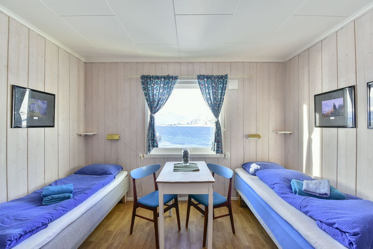 Eine Hütte mit zwei Betten, einem kleinen Tisch, Stühlen, einem fenster mit Vorhängen und gerahmten Bildern an den Wänden.