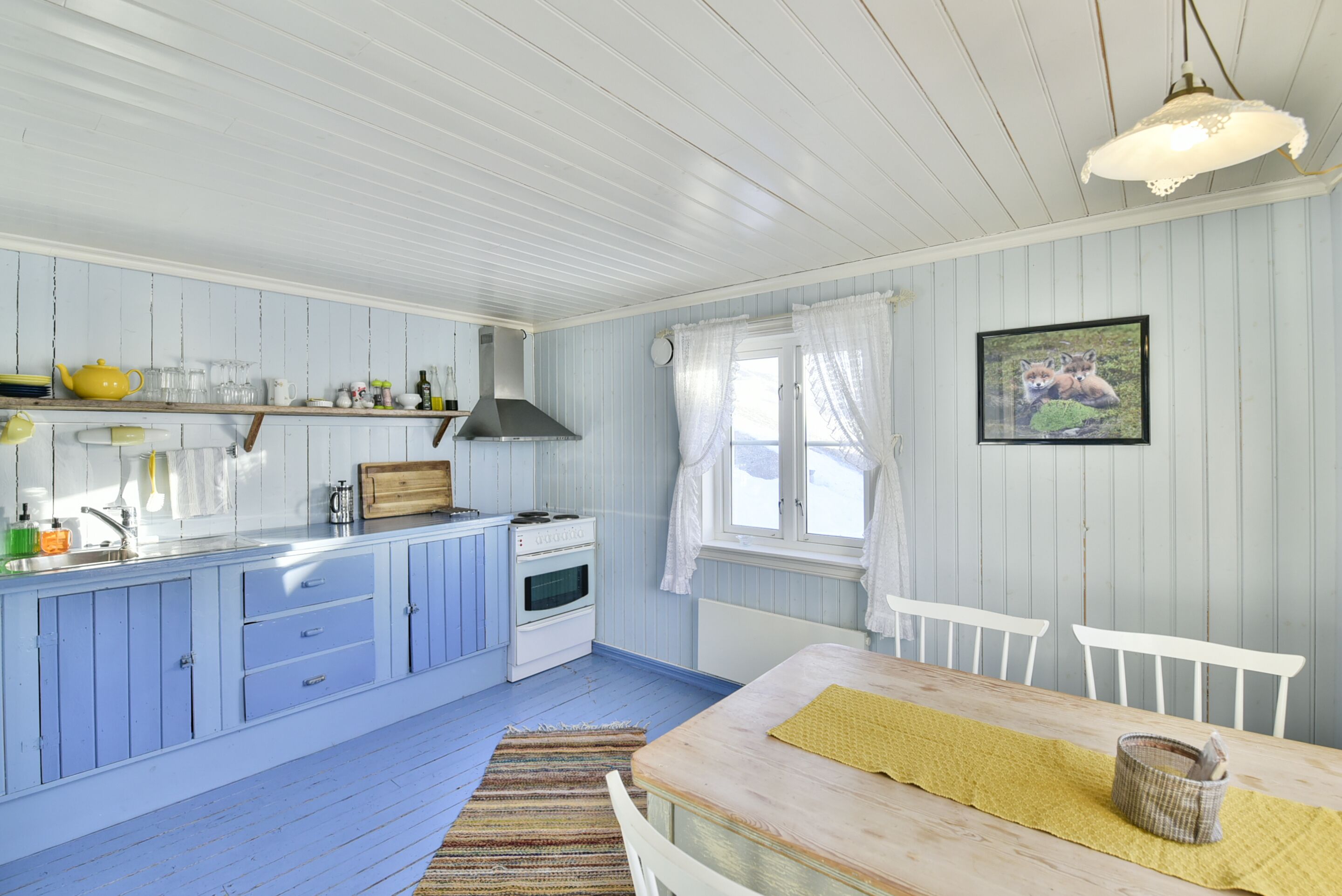 A kitchen with blue cabinets, a wooden table, and a window with curtains.