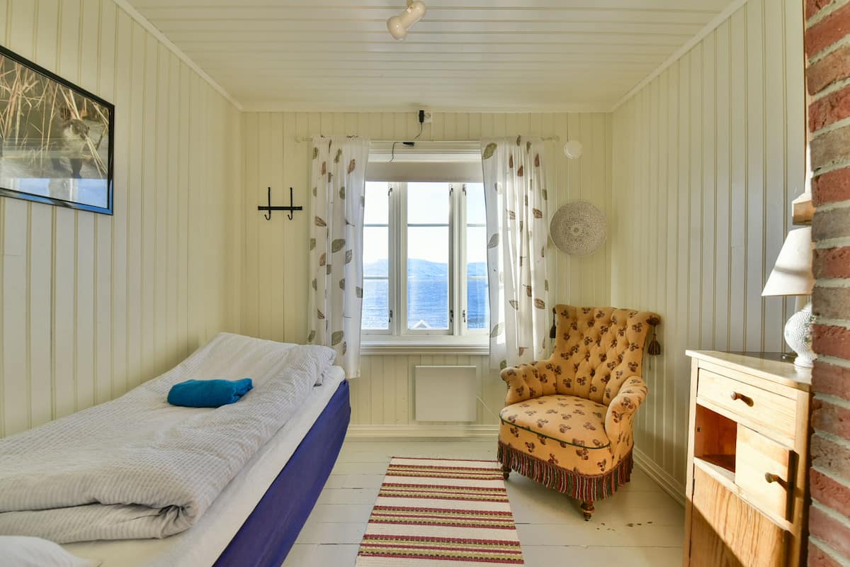 Ein Zimmer mit einem Bett, einem Sessel, einem Fenster mit Vorhängen, einem Heizkörper und einem an der Wand hängenden Bild.