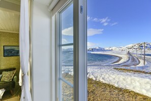 A room with a large window overlooking a snowy landscape and a winding road. - Kongsfjord Arctic Lodge (Berlevag)