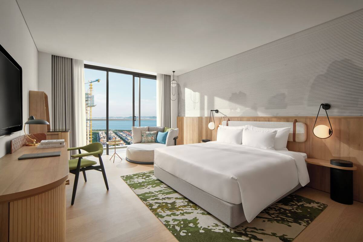 Habitación de hotel moderna con una cama grande, un escritorio con silla, televisión y vistas al océano a través de amplios ventanales.