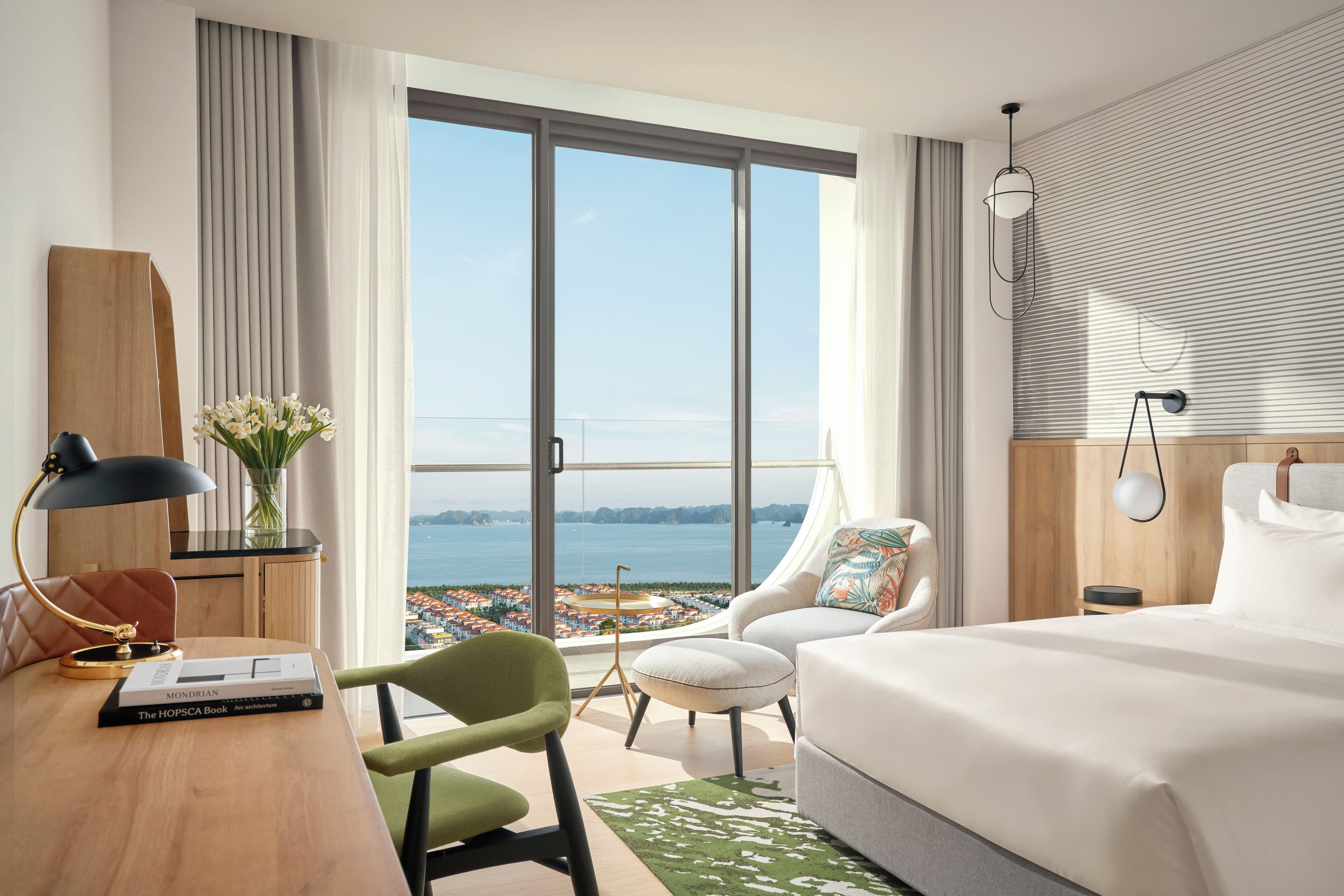 A modern hotel room with a large bed, a desk with a lamp, a chair, and a view of the sea through large windows.