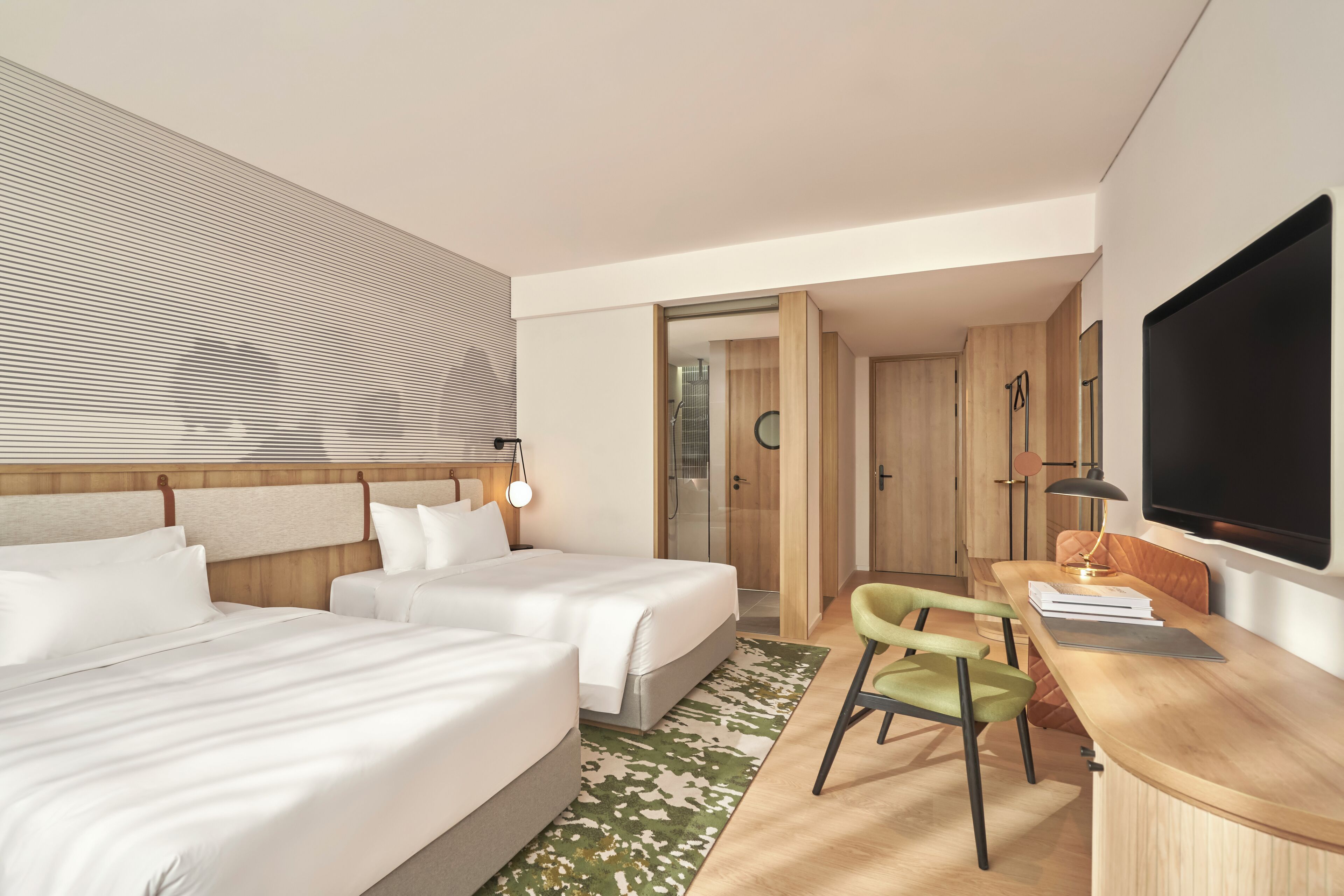 A hotel room with two beds, a desk with a chair, a TV, and a bathroom visible through an open door.