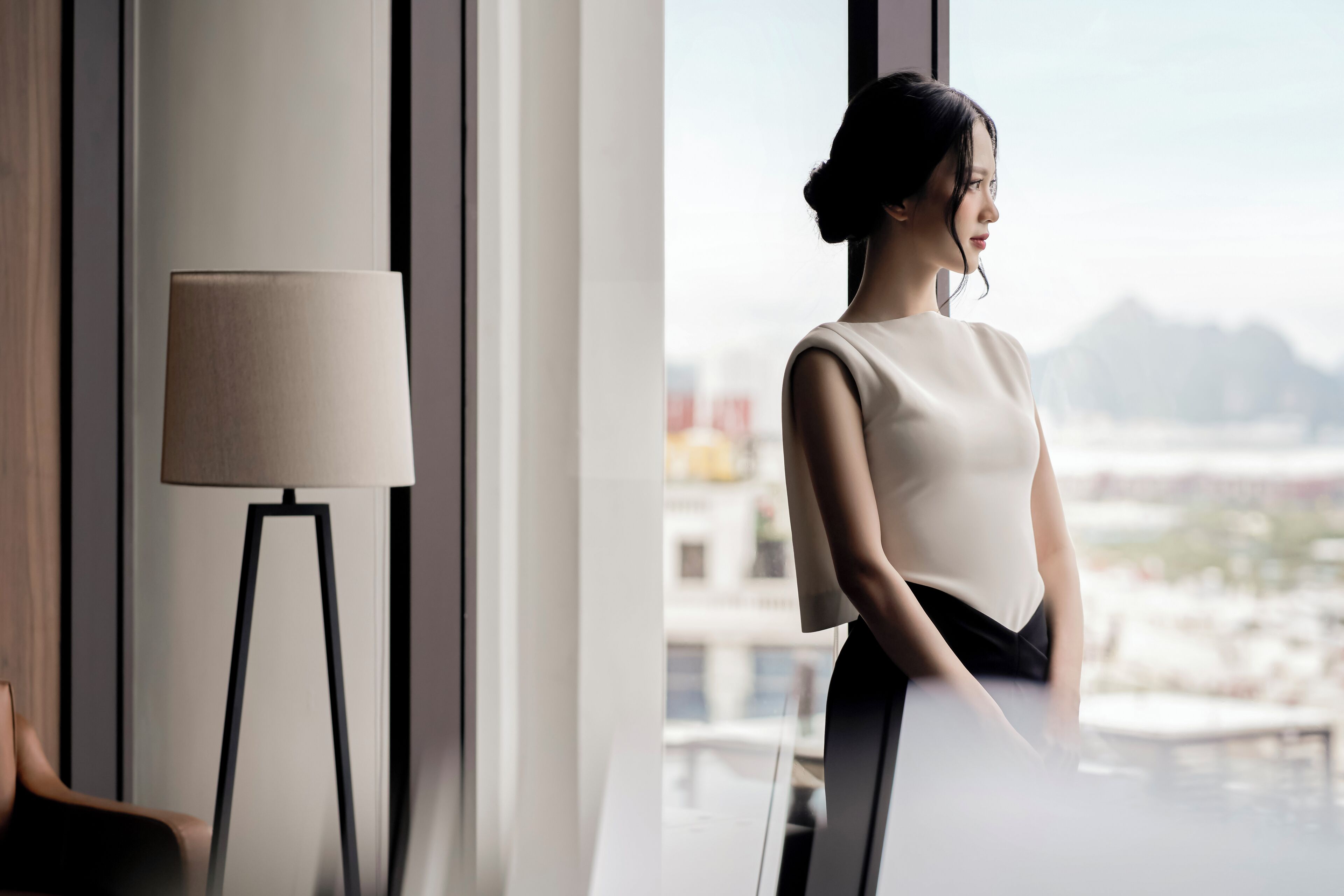 A modern hotel room with a large window, a standing lamp, and a view of the cityscape.