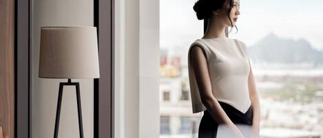 A modern hotel room with a large window, a standing lamp, and a view of the cityscape.